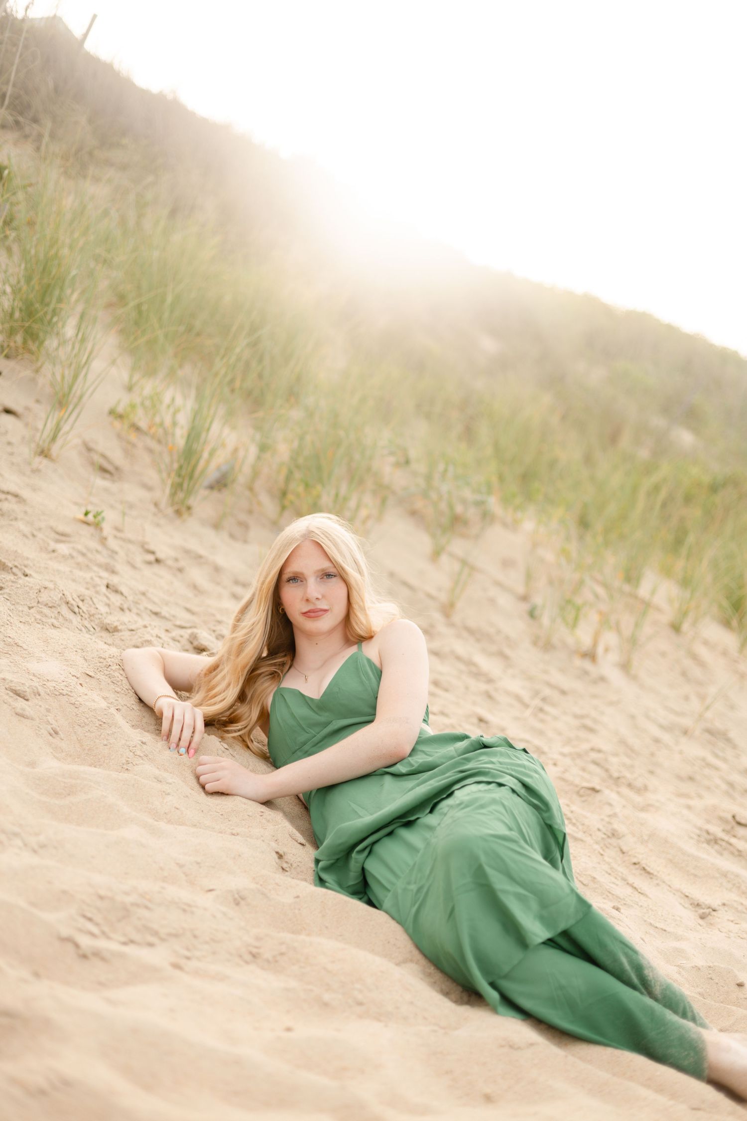 A woman in a green dress reclines gracefully on sandy dunes with grass and soft sunlight in the background.