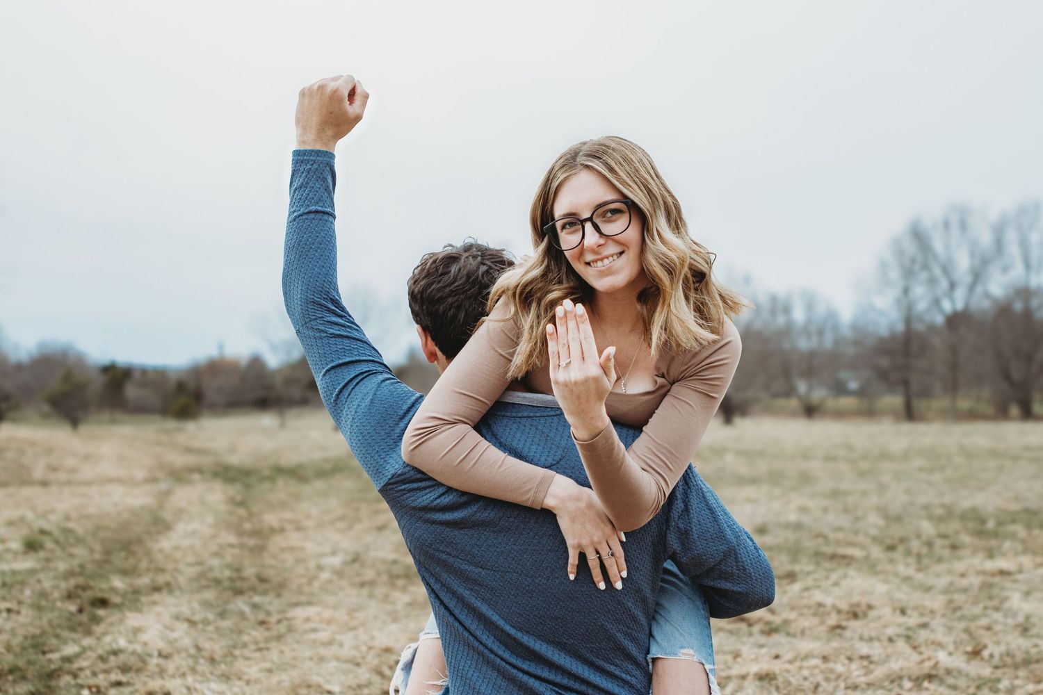 Couple celebrating engagement in a field, woman showing diamond ring while man raises fist in joy.