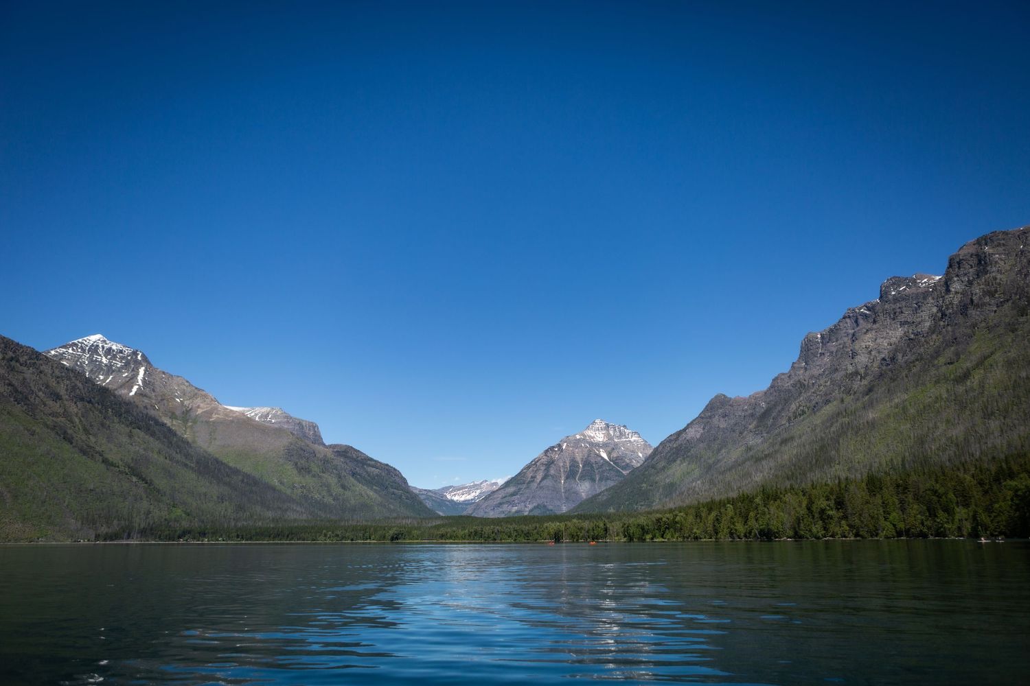 Serene mountain lake with snow-capped peaks, lush green forests, and a clear blue sky reflected in calm turquoise waters.