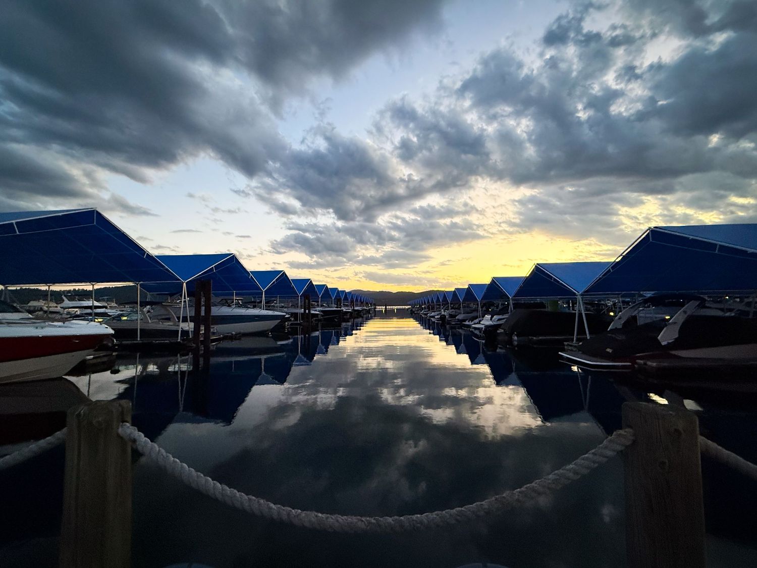 Marina at sunset with blue boat canopies reflecting on calm water under dramatic cloudy sky.