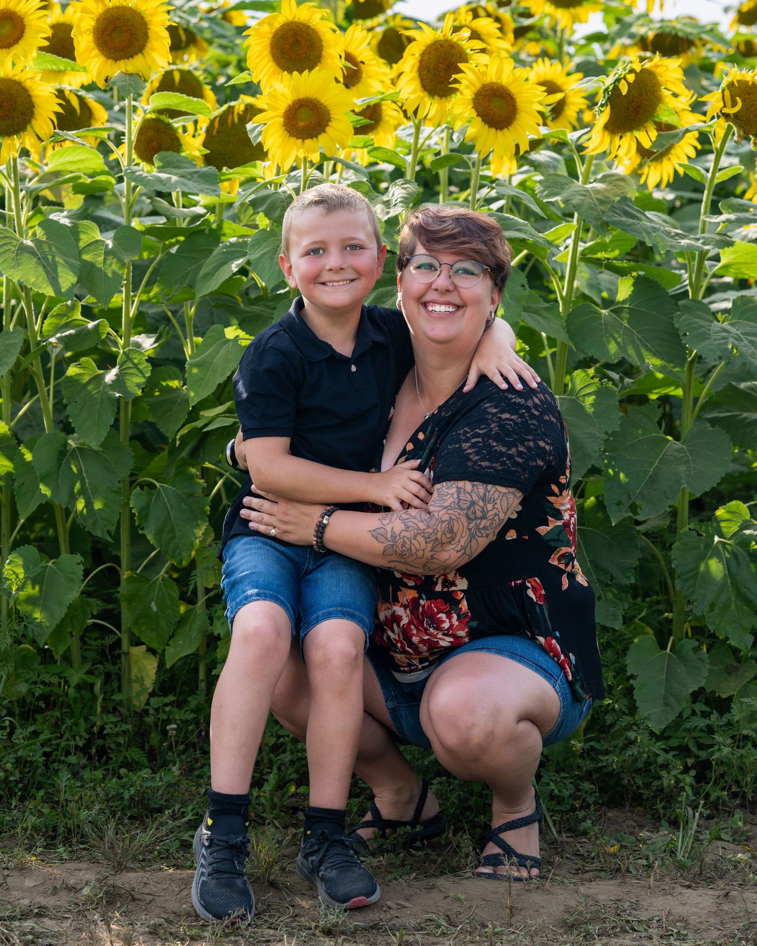 A woman and young boy smile together in a sunflower field, the boy sitting on her knee in a warm outdoor portrait.