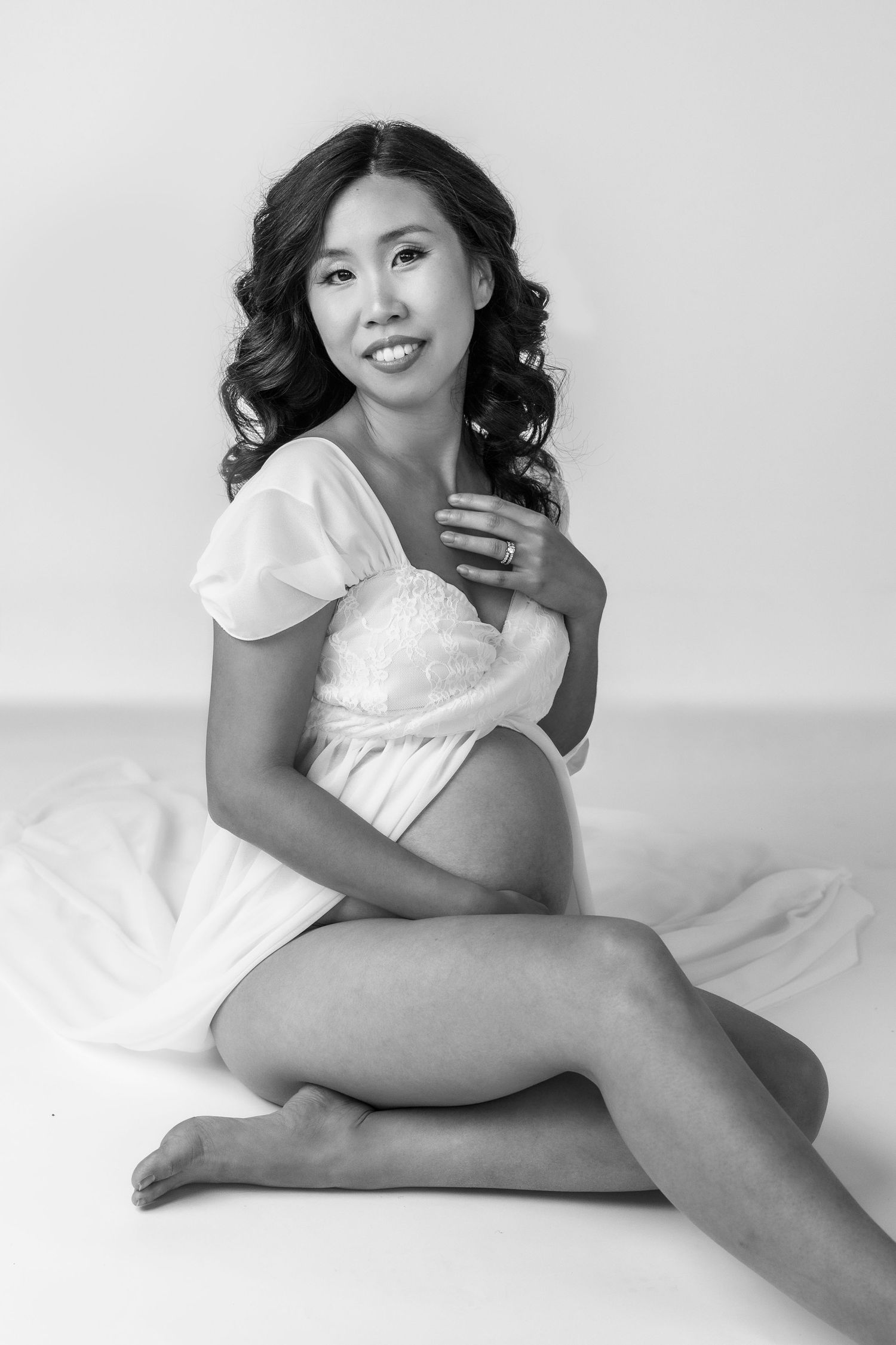 Black and white maternity photo of a smiling pregnant woman in white dress sitting gracefully on white fabric.