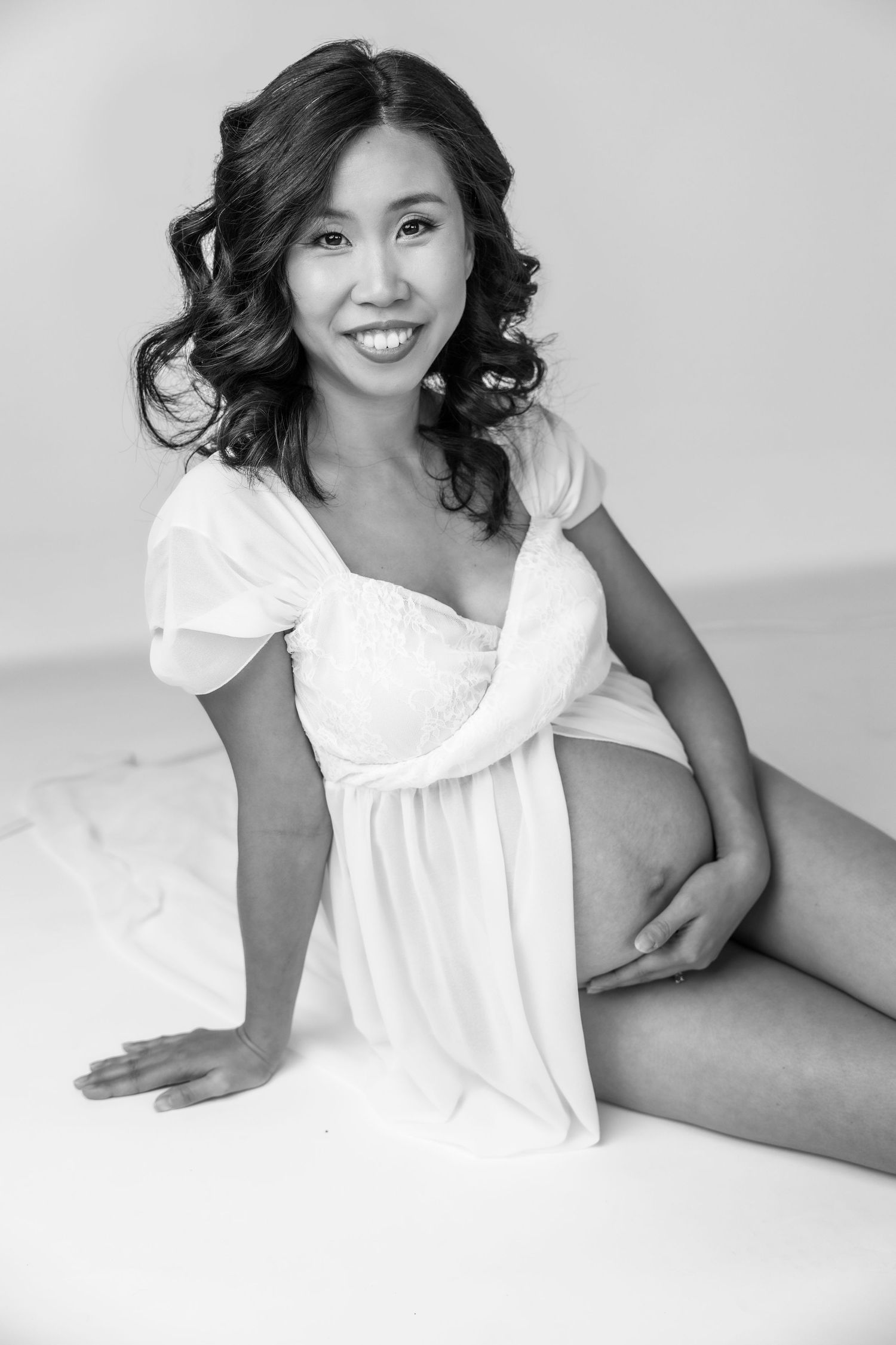 Black and white portrait of a smiling pregnant woman in a white dress sitting gracefully on a white surface.
