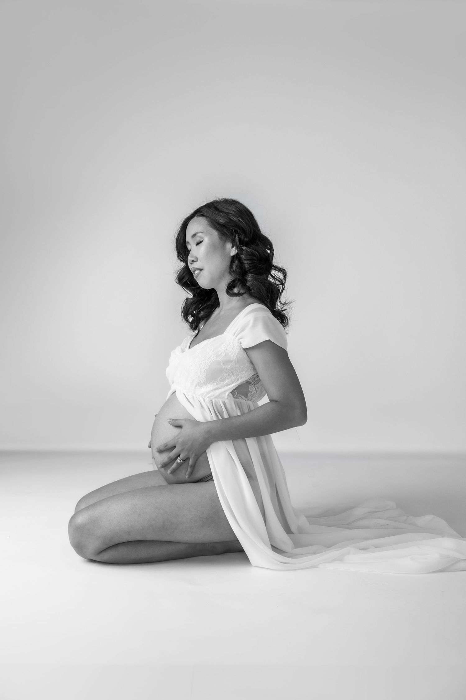 Black and white maternity photo of a pregnant woman in a white dress kneeling gracefully on a studio floor.
