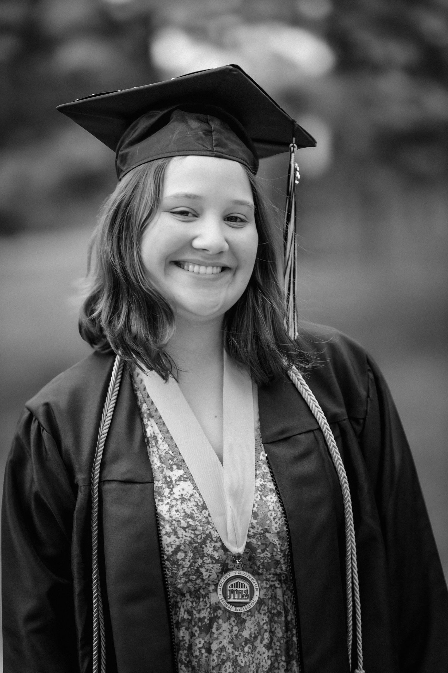 Smiling young woman in graduation cap and gown with honor cords and medal, black and white portrait photo.
