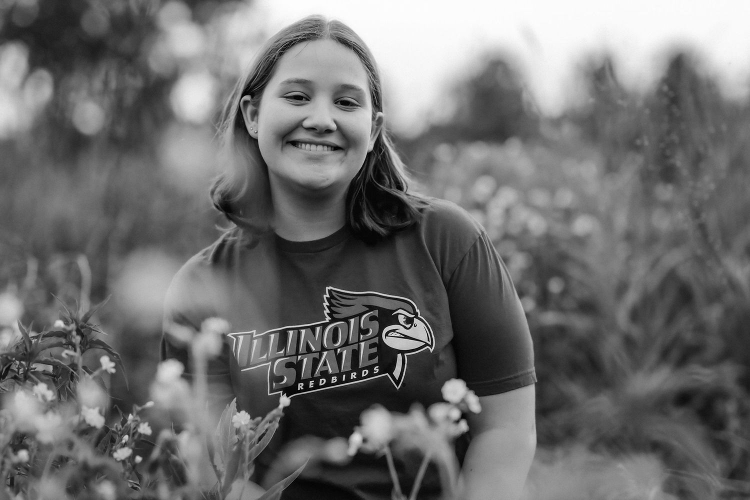 Smiling young woman wearing an Illinois State University t-shirt standing outdoors among wildflowers in black and white.