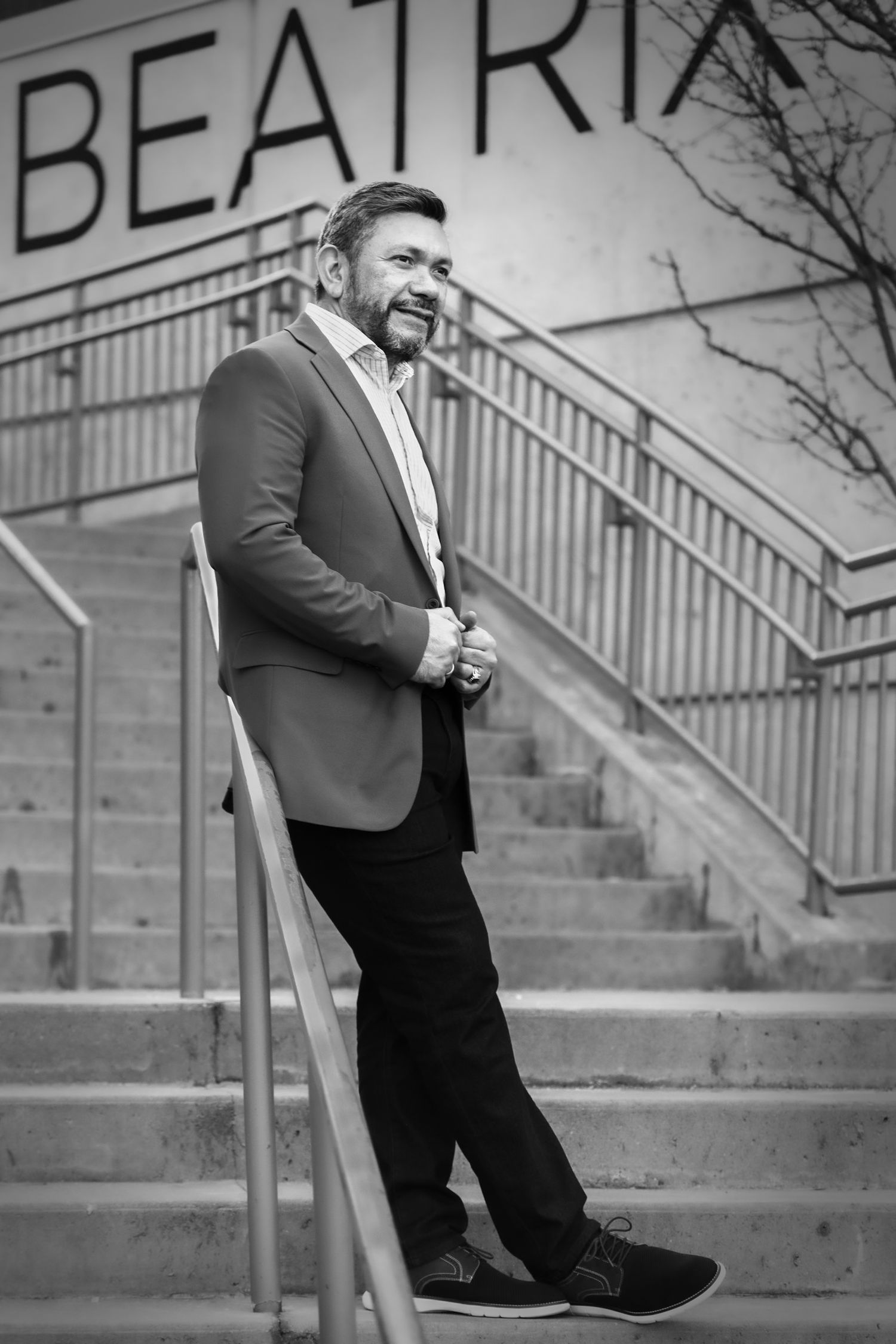 Black and white photo of a smiling man in a blazer leaning on a staircase railing outdoors near a Beatrix sign.