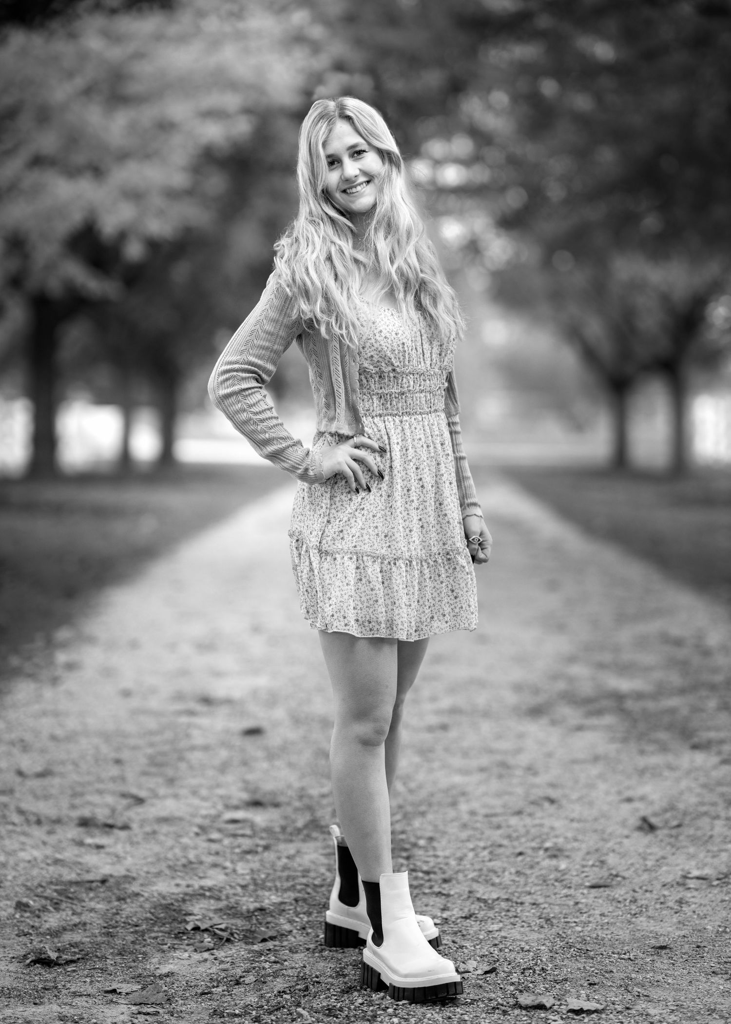 Black and white photo of a smiling blonde woman in a floral dress and boots standing on a tree-lined path.