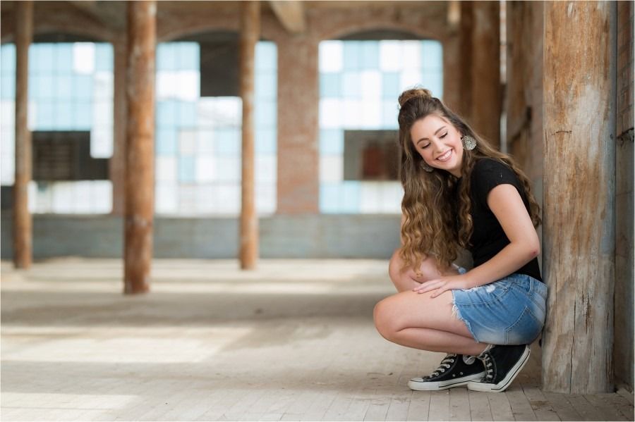 Smiling teenage girl crouching against a wooden pillar in an industrial space, wearing denim shorts and black sneakers.