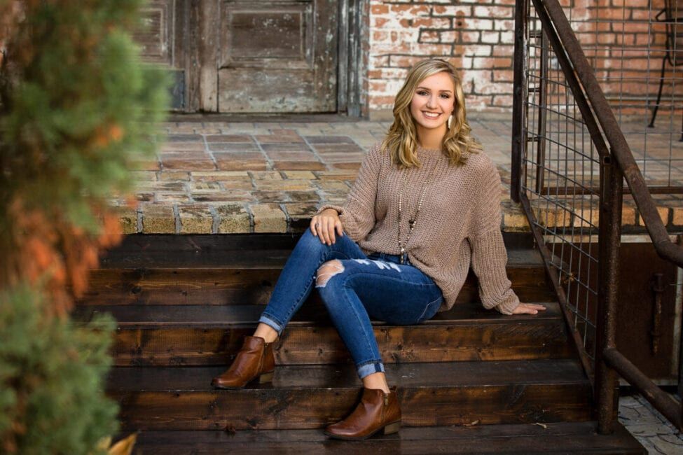 Young woman smiling in ripped jeans, brown sweater, and ankle boots, sitting on wooden steps by a brick building.