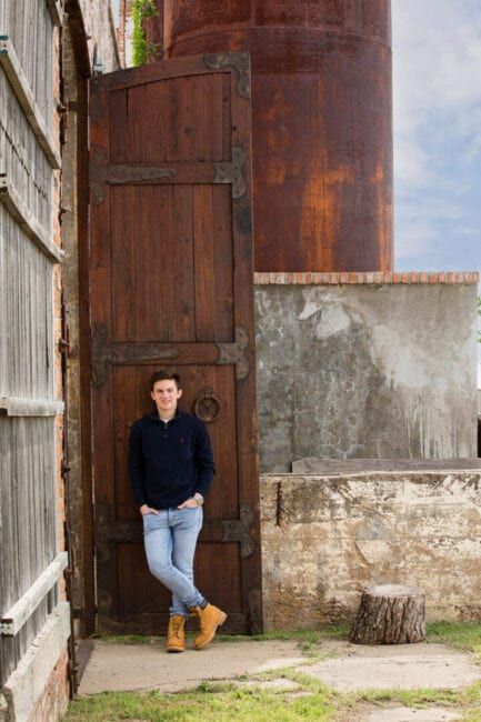 Young man in navy sweater and jeans leans against rustic wooden door near vintage silo outdoors.