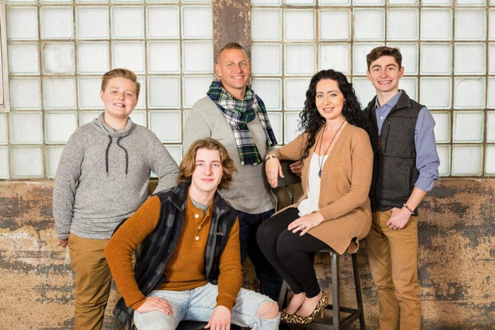 A smiling family of five poses together in a rustic studio setting for a professional portrait photo.