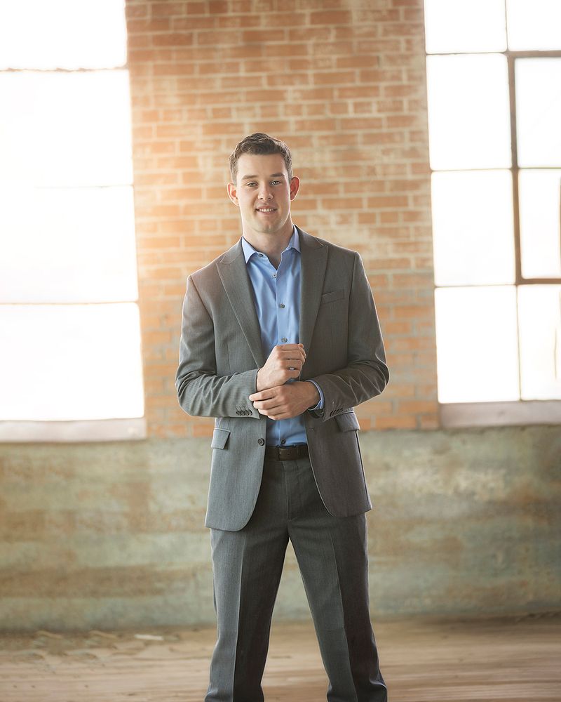 Professional man in gray suit and blue shirt smiling in a bright brick-walled room.