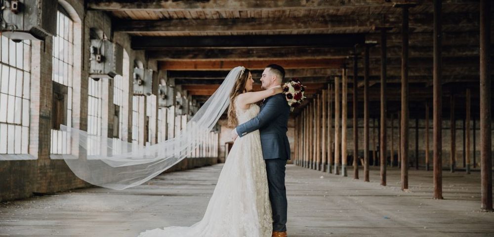 Bride and groom share a romantic embrace in a rustic industrial venue, her long veil flowing gracefully behind her.