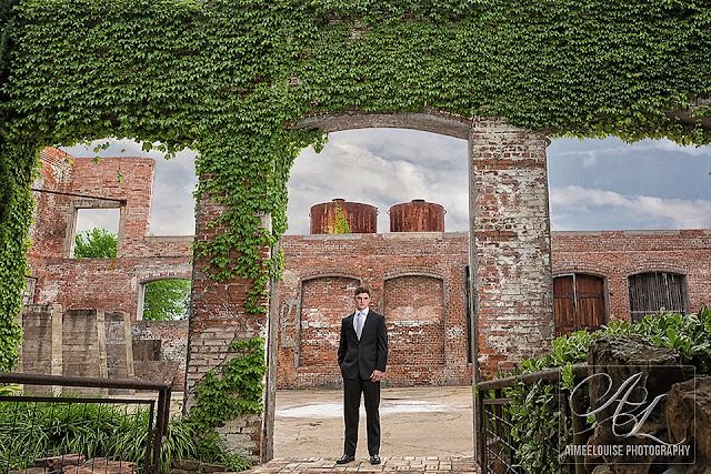 Groom in black suit poses in ivy-covered brick archway at rustic outdoor wedding venue.