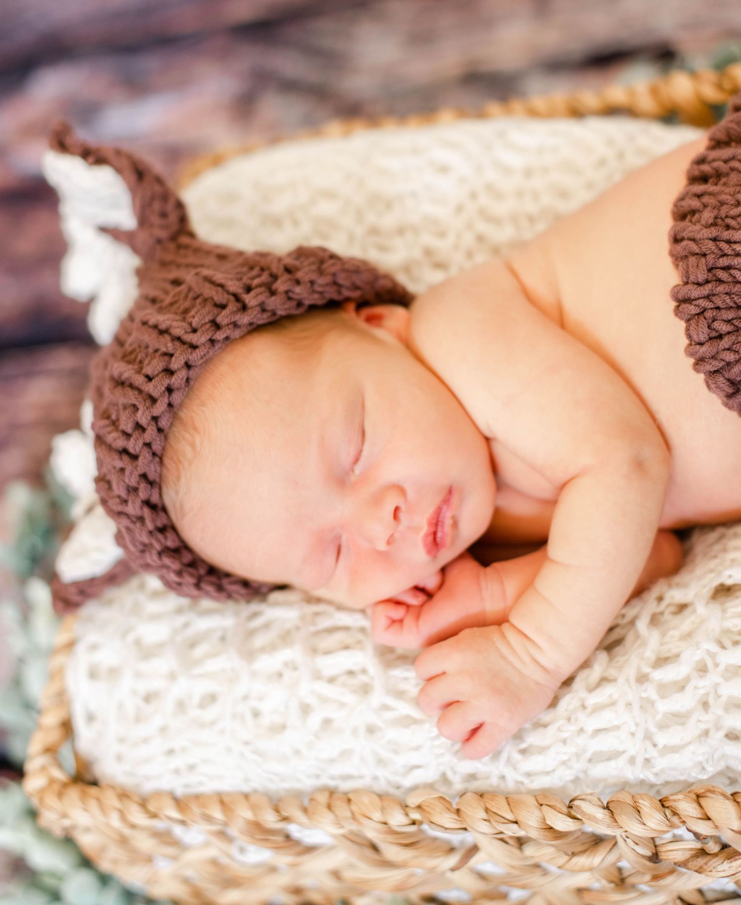 Sleeping newborn baby in brown knit bear hat resting peacefully in a woven basket on white blanket.