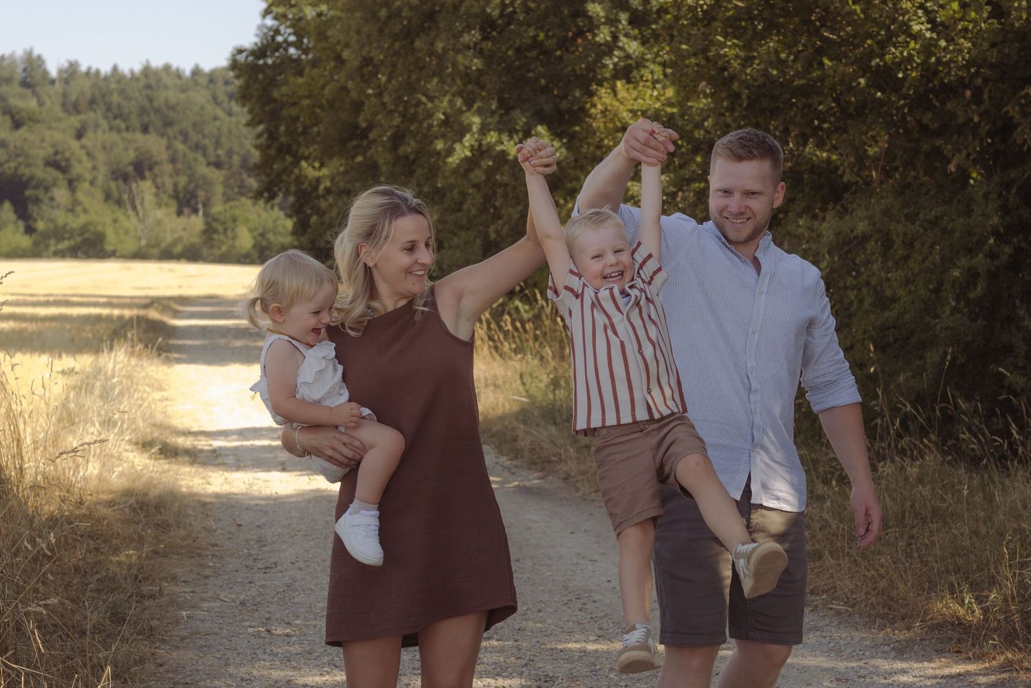 Glückliche Familie mit zwei Kindern bei einem natürlichen Familienshooting auf einem sonnigen Feldweg im Aargau.