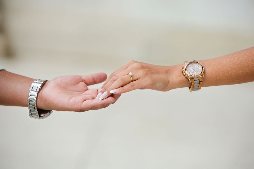 Two hands reaching toward each other, one wearing a diamond ring and gold watch, the other a silver watch.