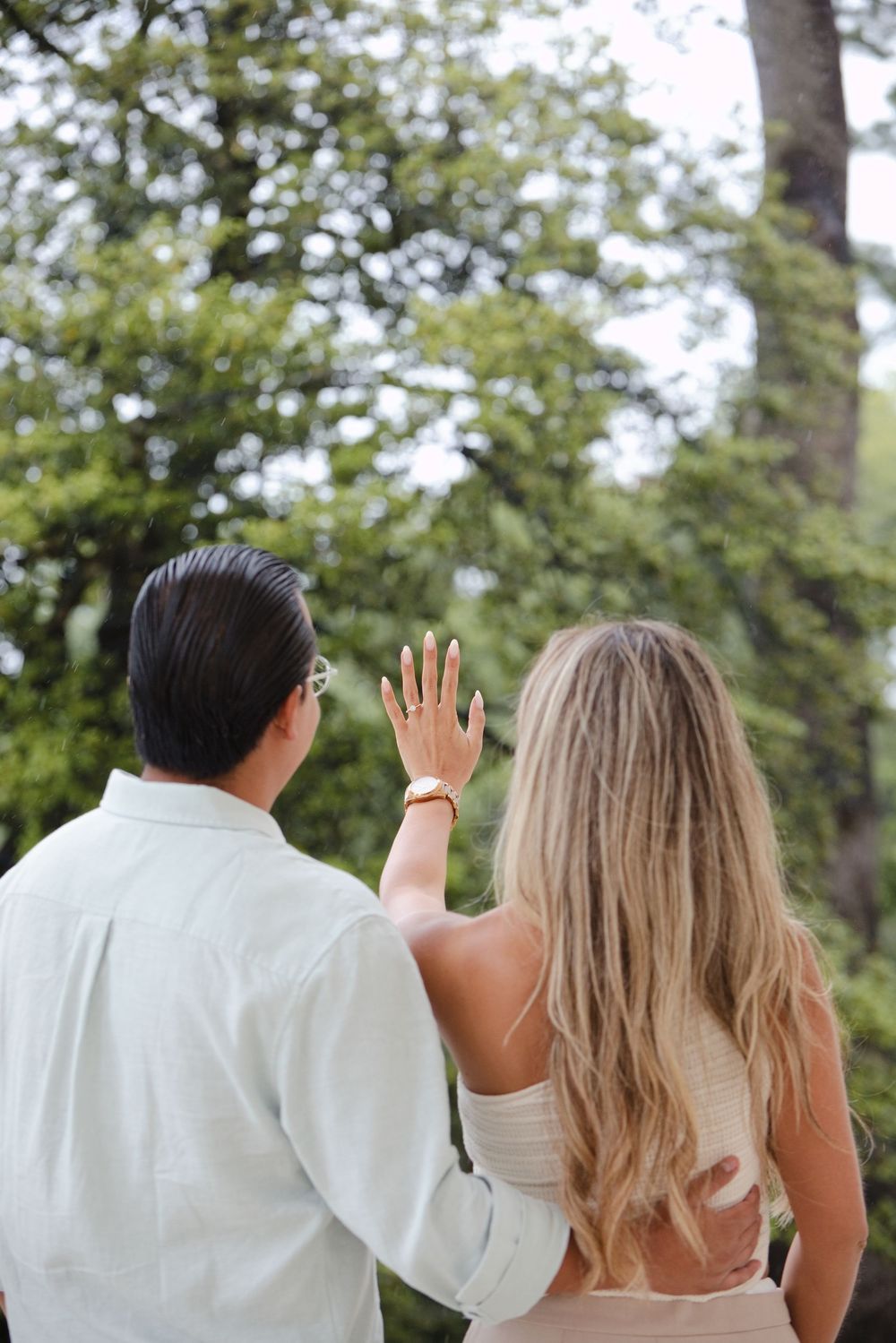 Couple standing outdoors facing trees, woman raising hand showing engagement ring, wearing watch and light clothing.