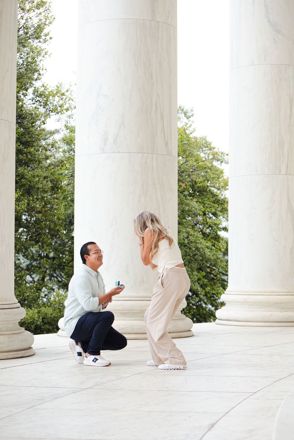 Man kneeling on one knee proposing to woman with ring box at Jefferson Memorial columns.