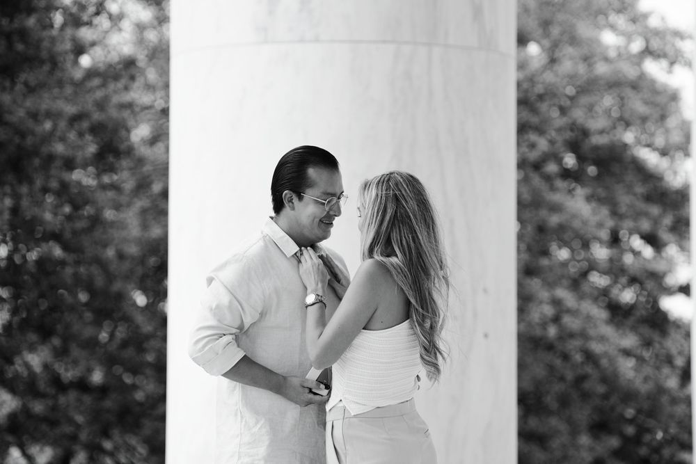 Black and white photo of a couple holding hands and gazing at each other beside a marble column outdoors.