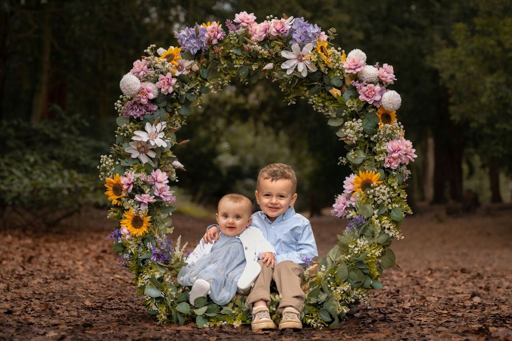 Two young children sit inside a large floral wreath with colorful flowers outdoors.