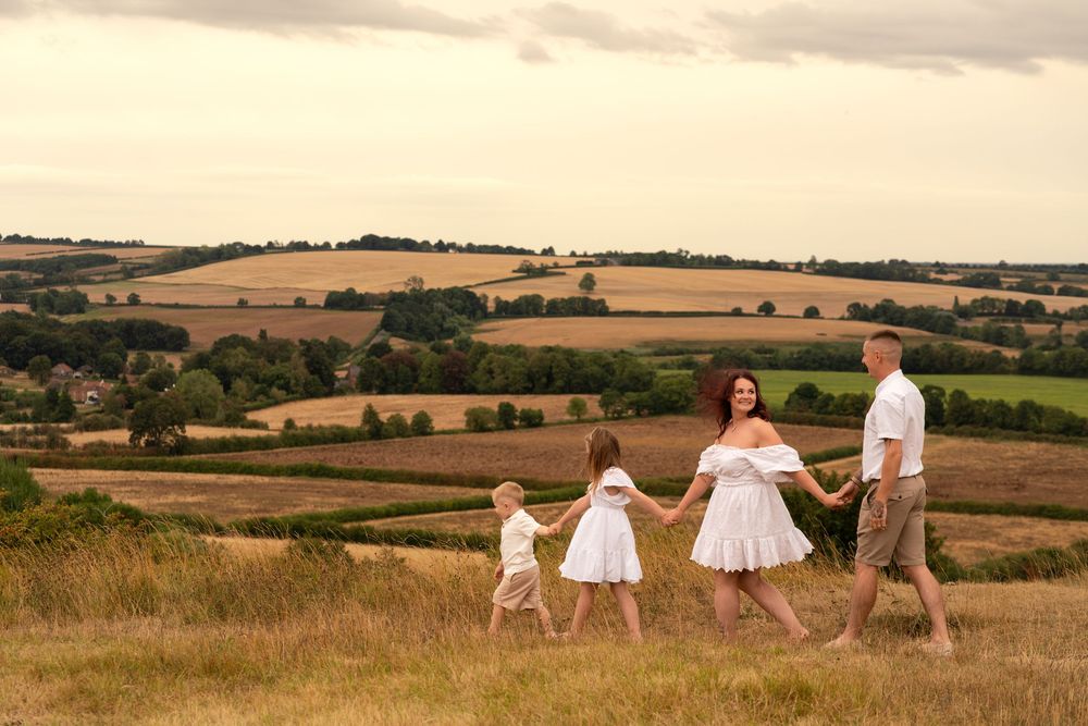 Family of four holding hands walking through golden countryside fields under a cloudy sky in rural England.