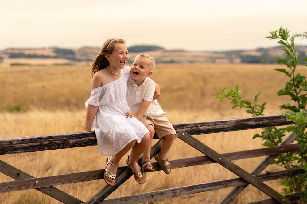 Two children sitting on a wooden fence laughing in a golden field, girl in white dress and boy in beige outfit.