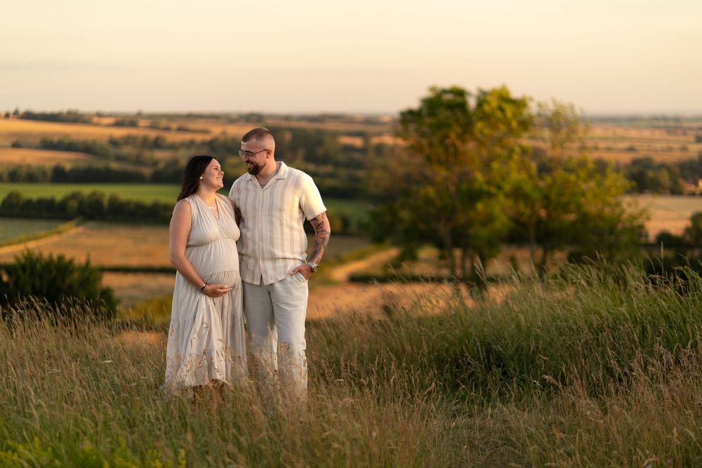 Pregnant couple standing in a golden field at sunset, sharing a tender moment with scenic countryside views.