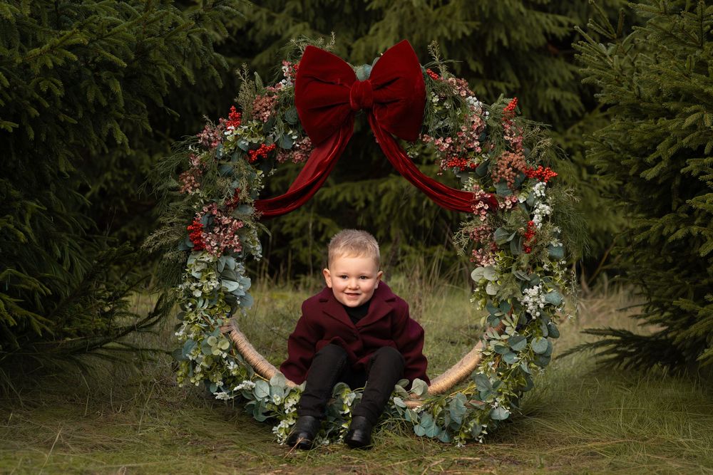 Young boy in burgundy outfit sits smiling inside a festive Christmas wreath with red bow outdoors.