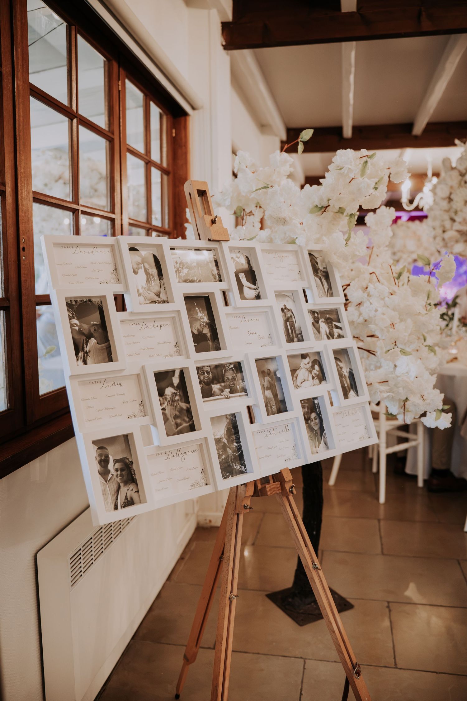 Elegant wedding photo display board with white frames on wooden easel, featuring black and white photos at a reception venue.