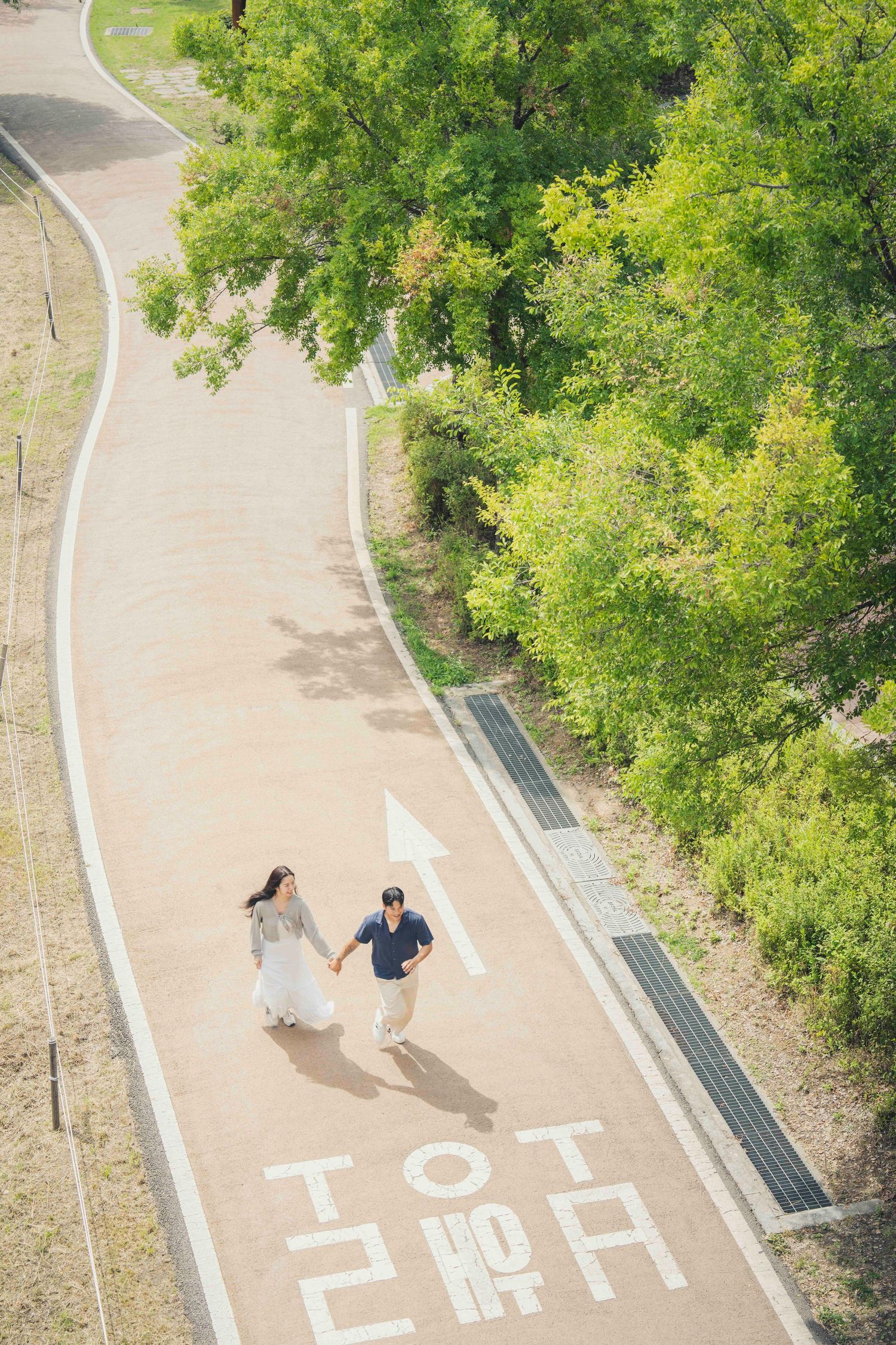 Aerial view of a couple holding hands walking on a Korean park path surrounded by lush green trees.