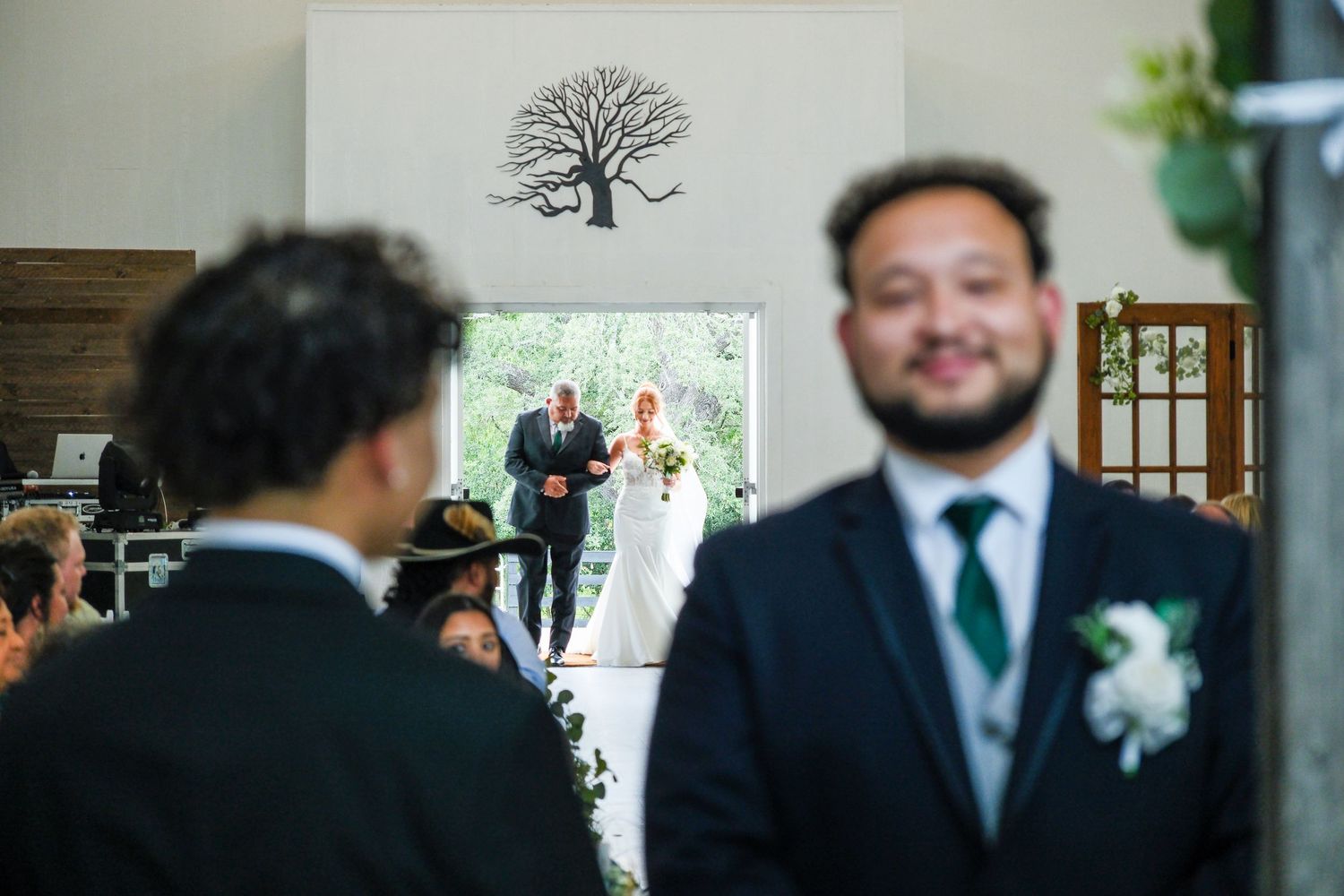 Groom smiling in foreground as bride walks down aisle with father in sunlit wedding venue with tree wall art.