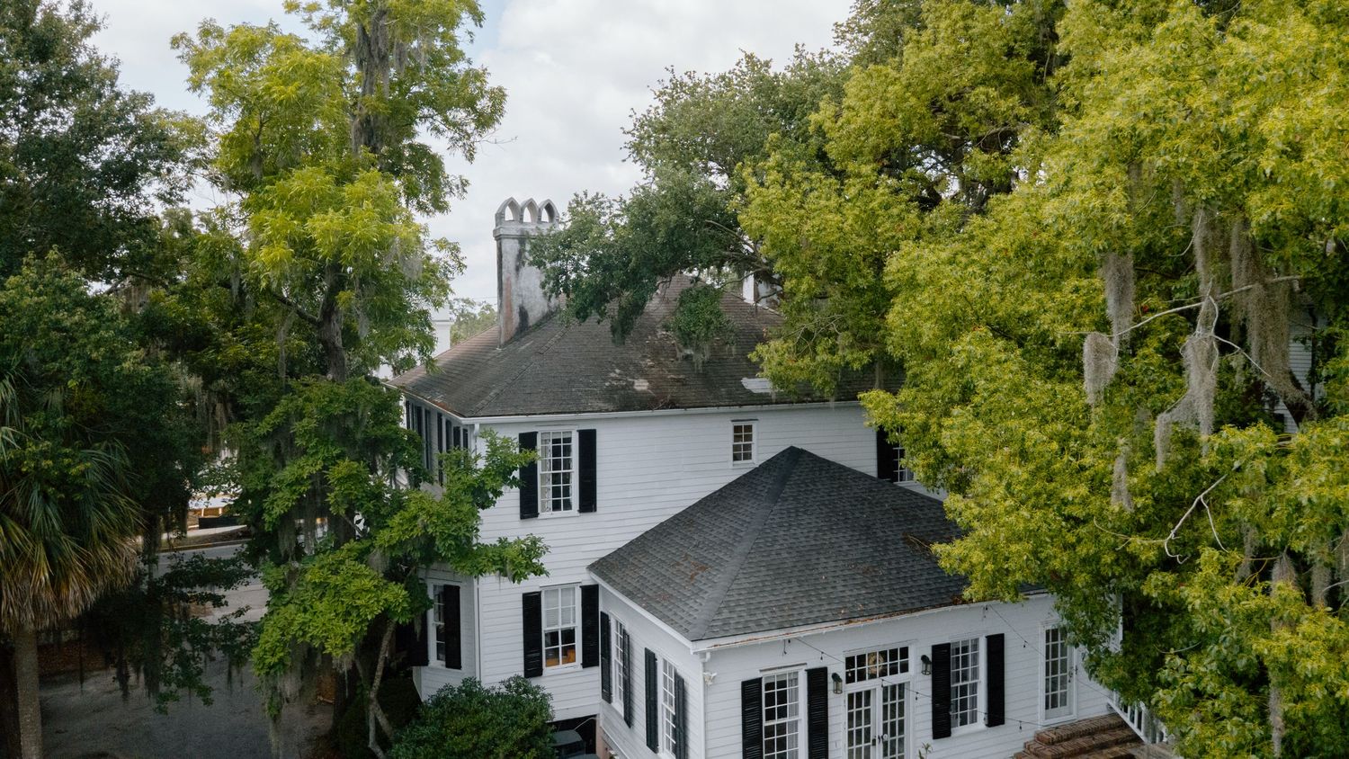 Aerial view of a white two-story historic Southern home surrounded by lush green oak trees with Spanish moss.