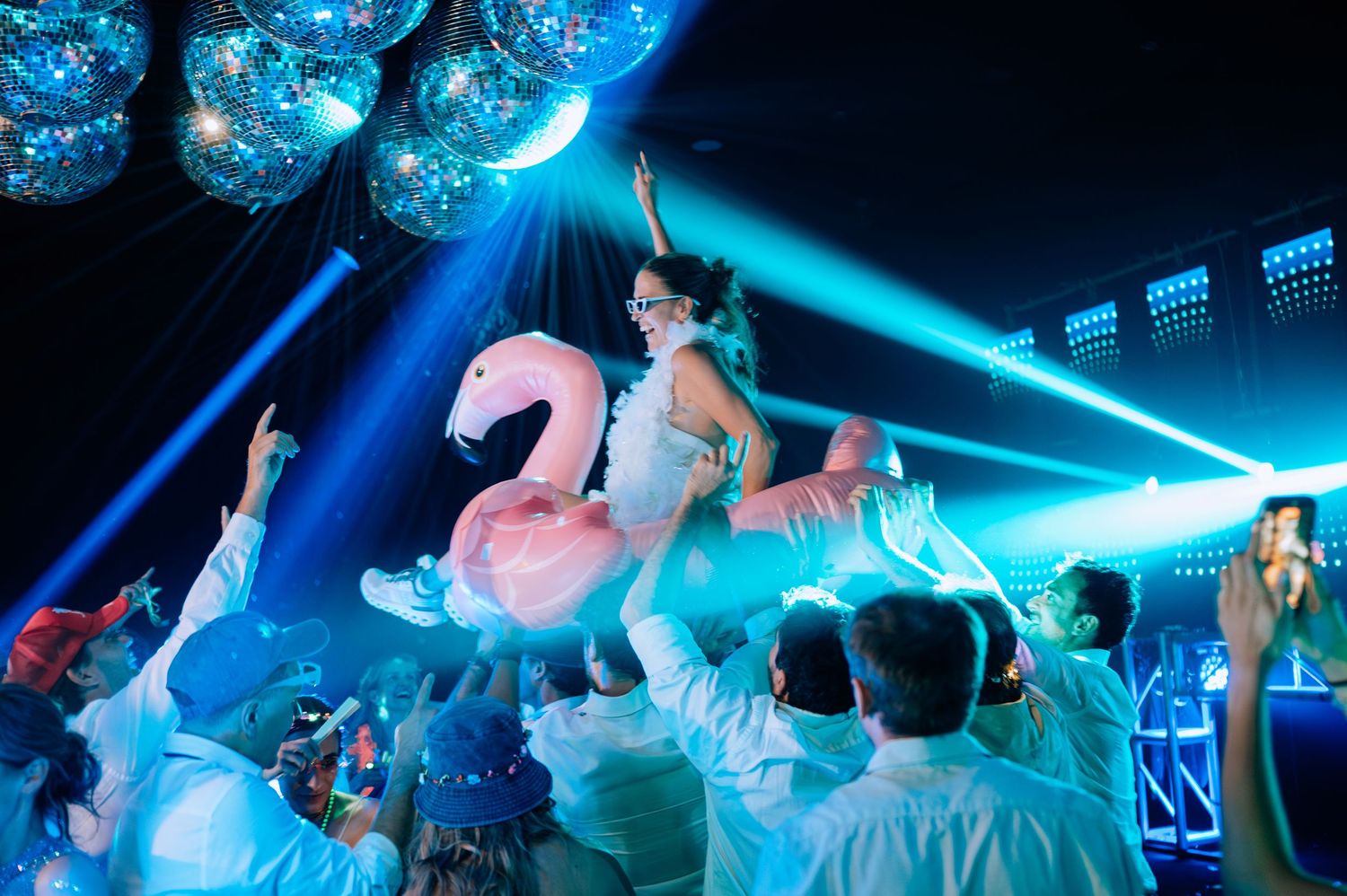 Joyful wedding guest lifted on pink flamingo float under disco balls at a vibrant reception party.
