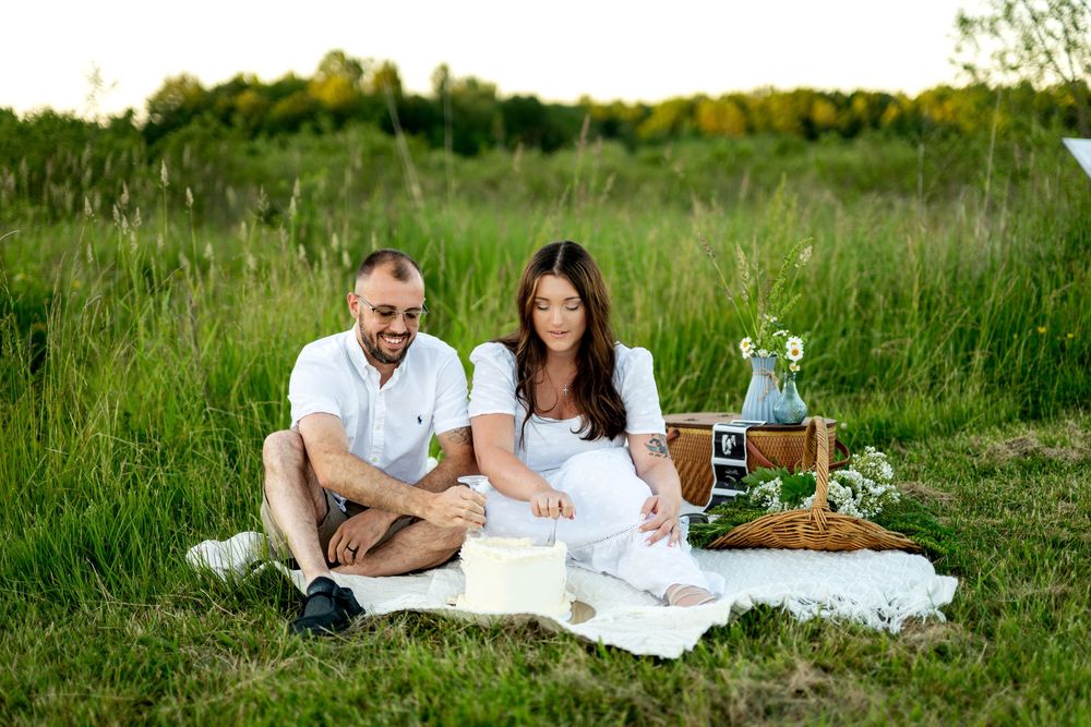 Couple sitting on white blanket in green field for a romantic outdoor picnic with wicker basket and flowers.
