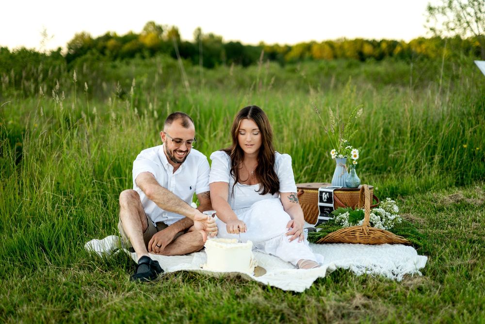 Couple in white outfits enjoying a romantic picnic on a blanket in a lush green field with a wicker basket.