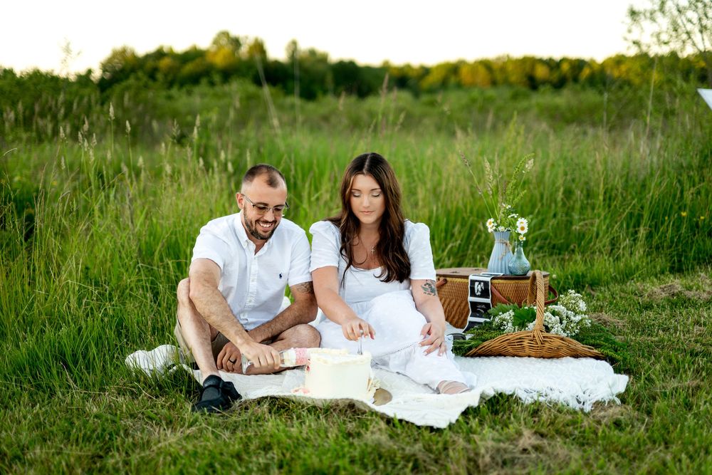 Pregnant couple enjoying a picnic on a white blanket in a green field, looking at ultrasound photos together.