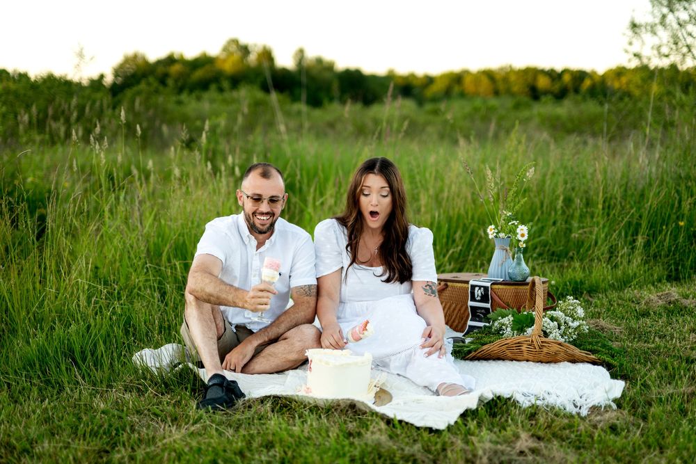 Couple sitting on white blanket in green field having a picnic, man holding sparkler, pregnant woman in white dress.