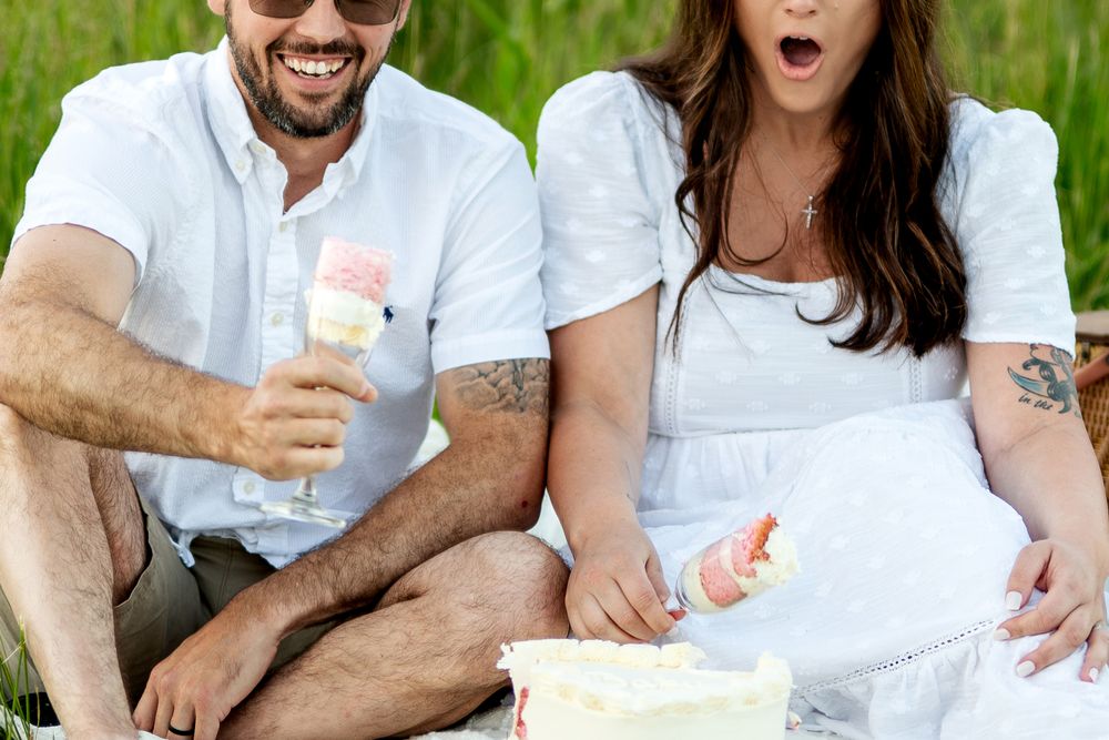 Couple sitting outdoors in white outfits sharing ice cream cones and white cake at a summer picnic.