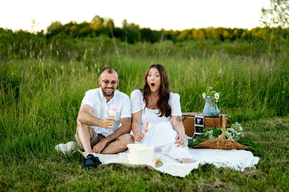 Surprised couple sitting on white blanket at outdoor picnic in green field, holding champagne glasses.