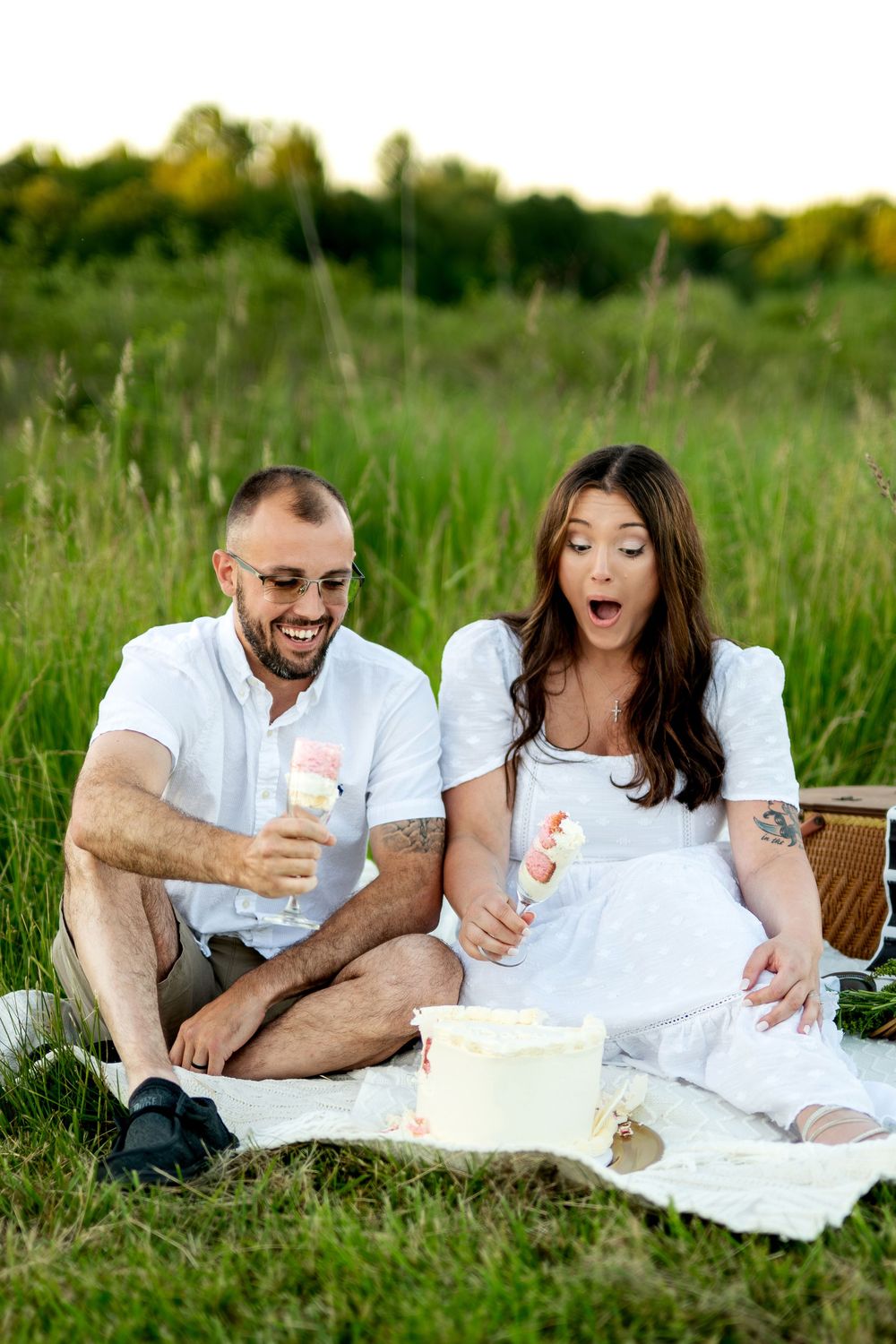 Couple enjoying a picnic in a green field, sharing cake and ice cream, woman in white dress appears surprised.