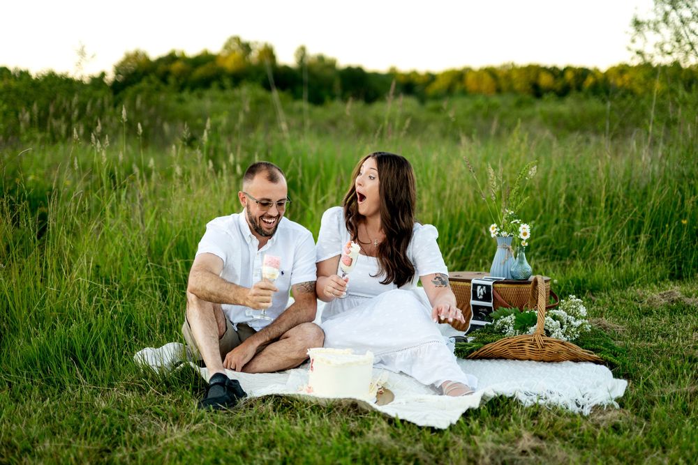Happy couple laughing together on white blanket during outdoor picnic in lush green field.