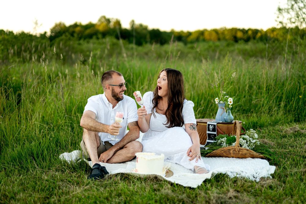 Couple laughing on white picnic blanket in green field, woman in white dress, man in white shirt, with basket and flowers.