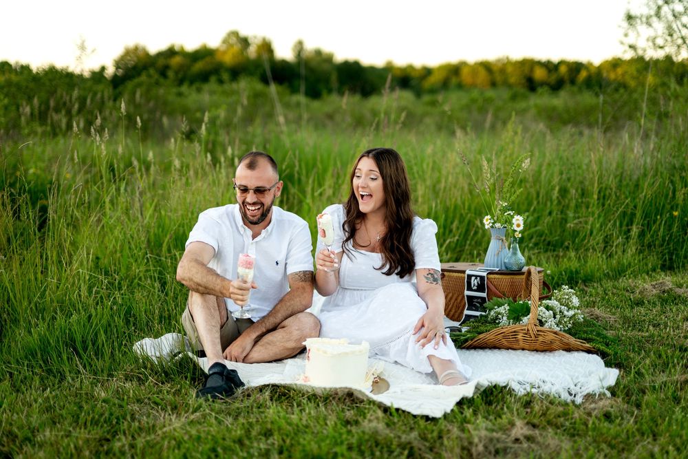 Couple laughing on white blanket at outdoor picnic in green field, holding champagne glasses with picnic basket nearby.