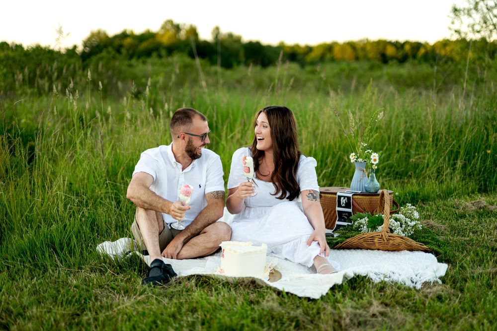 Happy couple sitting on white blanket in sunny field, sharing a romantic picnic with champagne and flowers.