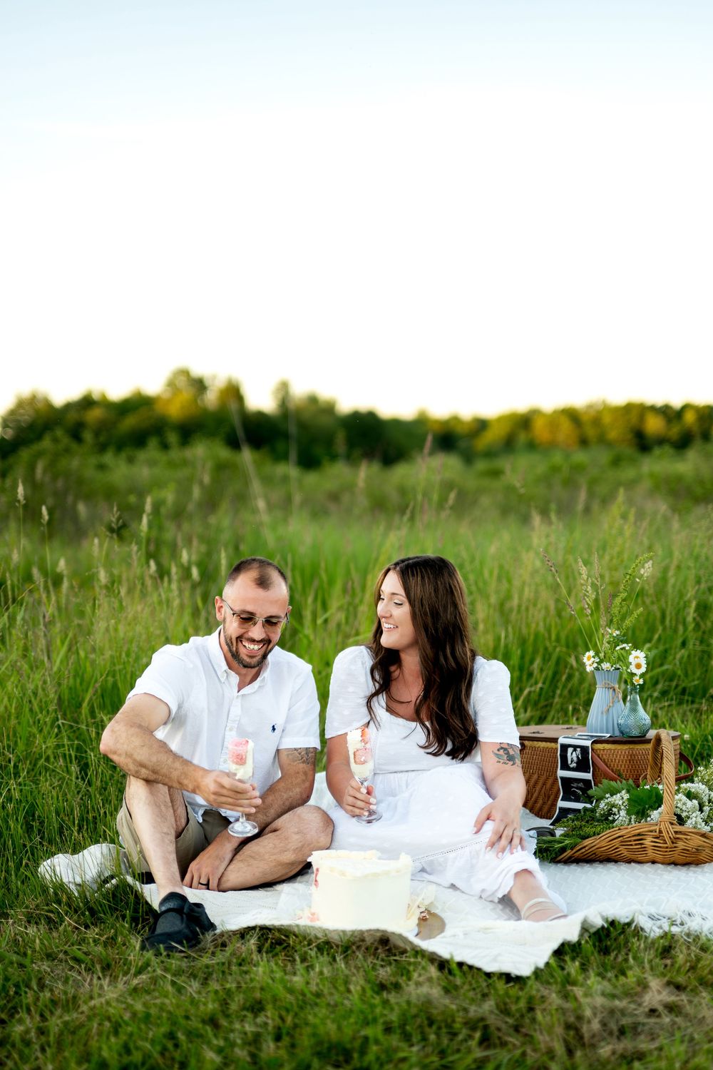 Couple in white outfits enjoying a romantic picnic in a green field, holding champagne glasses and smiling at each other.