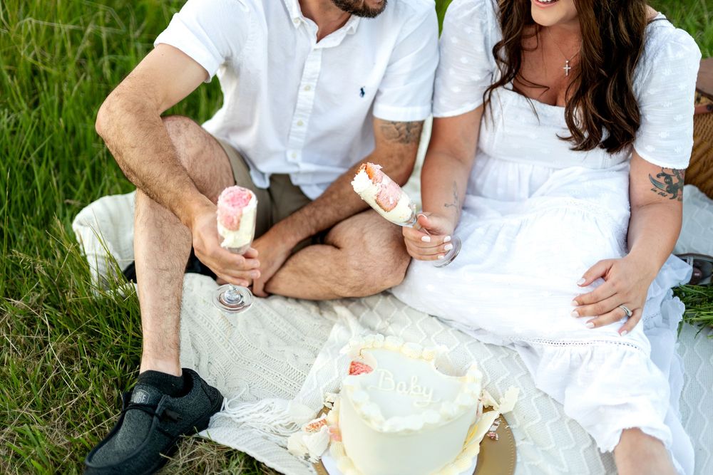 Couple in white outfits sitting on blanket outdoors, sharing popsicles near a white cake.
