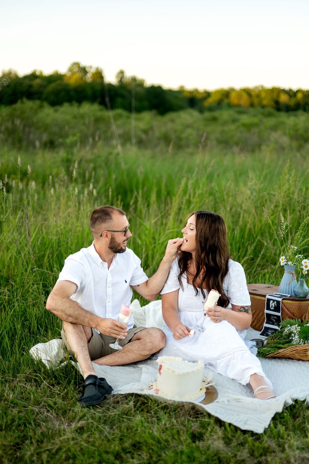 Couple enjoying a romantic picnic in a green field, man feeding woman while holding ultrasound photos nearby.