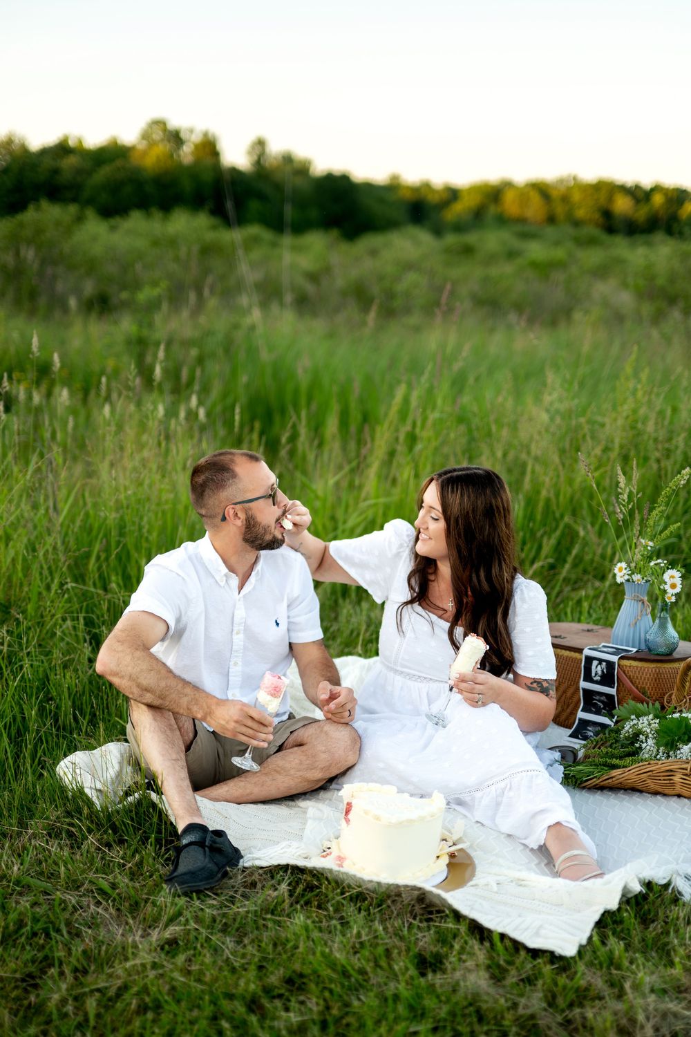 Happy couple sharing a tender moment at an outdoor picnic, woman feeding man while sitting on white blanket in green field.